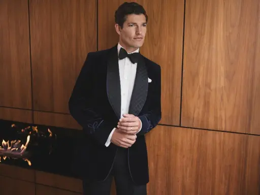 A man in a sophisticated navy velvet tuxedo, embodying perfect black tie attire, stands by a modern fireplace.