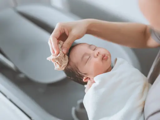 Bain d'un nouveau-né : une main nettoie délicatement la tête du bébé avec une petite serviette. Une scène paisible qui rappelle la liste produits toilette bébé naissance.