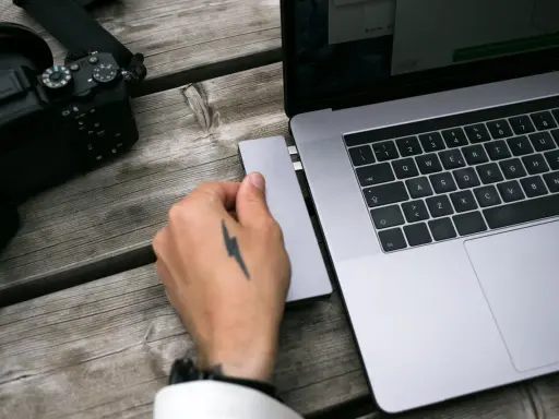 Dłoń z tatuażem pioruna podłącza adapter do laptopa, obok leży aparat. Idealny zestaw, jaki laptop do obróbki zdjęć?