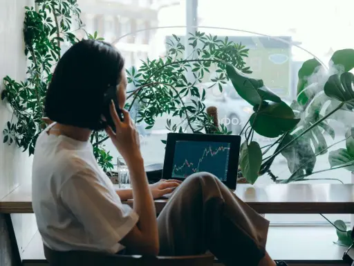 Kobieta w białej koszulce rozmawia przez telefon, pracując na laptopie z wykresem giełdowym. To jej wolny zawód, który pozwala jej pracować w otoczeniu zieleni.