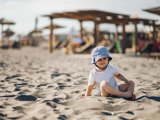 Uroczy maluch w pasiastym kapelusiku siedzi na piaszczystej plaży. Idealne miejsce, gdzie na wakacje z rocznym dzieckiem.