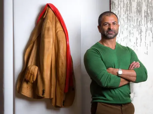 Livio De Marchi, artiste sculpteur, pose à côté de son manteau en bois sculpté, une œuvre saisissante.