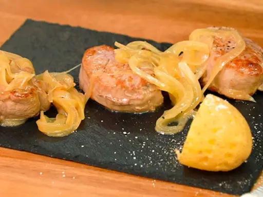 Tiernos medallones de carne al jerez, cubiertos con cebolla caramelizada y acompañados de una porción de patata.