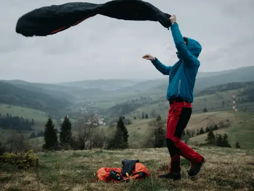 Osoba w niebieskiej kurtce i czerwonych spodniach rozkłada śpiwór puchowy czy syntetyczny na tle górskiego krajobrazu.