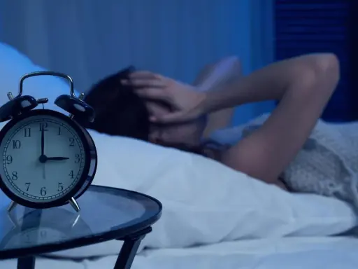 Eine Person liegt im Bett, die Hände über dem Gesicht, während ein Wecker auf 4 Uhr steht. Warum wache ich immer um 4 Uhr auf?