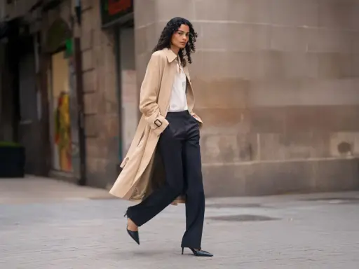 A woman walks confidently in dark palazzo pants and a trench coat, proving they are still in style.