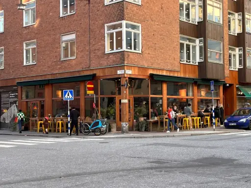 Ett mysigt hörnkafé i **Södermalm, Stockholm** med folk som sitter vid gula stolar och bord utomhus. En blå cykel med barnvagn står parkerad.