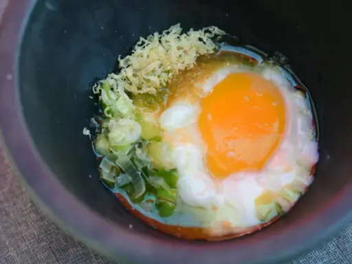 Ein perfekt zubereitetes Onsen-Ei mit Frühlingszwiebeln und geriebenem Ingwer in einer dunklen Schüssel.