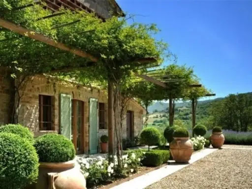 Splendide idee case di campagna esterni: un pergolato verde ombreggia una casa in pietra con persiane verdi, circondata da piante in vaso e un paesaggio collinare.