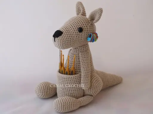 Canguro amigurumi beige sentado, con ganchillos en su bolsa y marcadores de puntos en su oreja.