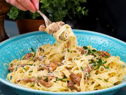 En gaffel lyfter en portion krämig pasta med kantareller, toppad med parmesan och färska örter i en vacker blå skål.