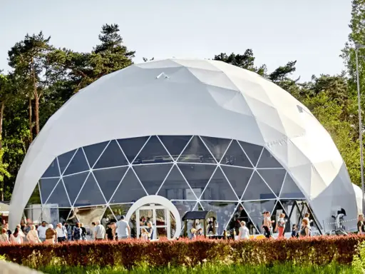 Una gran cúpula estructura blanca, con ventanas triangulares, se alza en un día soleado. Gente pasea frente a ella, rodeada de árboles verdes.