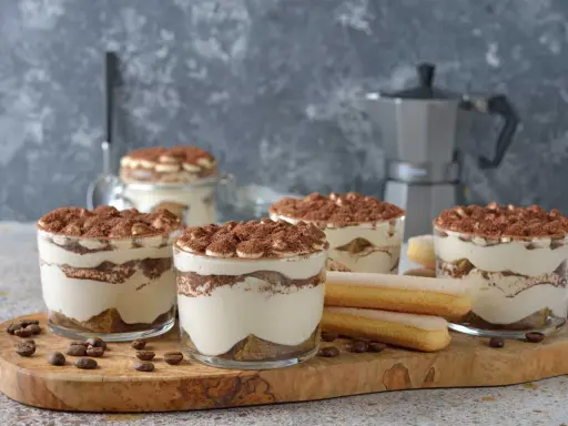 Plusieurs verrines de tiramisu sans café, garnies de cacao, côtoient des biscuits à la cuillère et des grains de café sur une planche en bois.