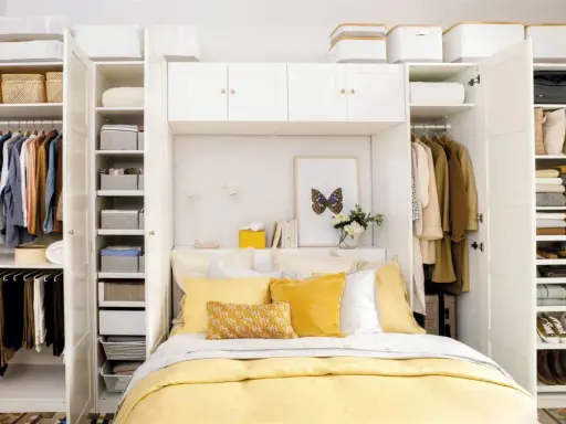 Habitación matrimonio pequeña con armario blanco, cama con ropa de cama amarilla y almohadas, y estanterías llenas de ropa y cestas.