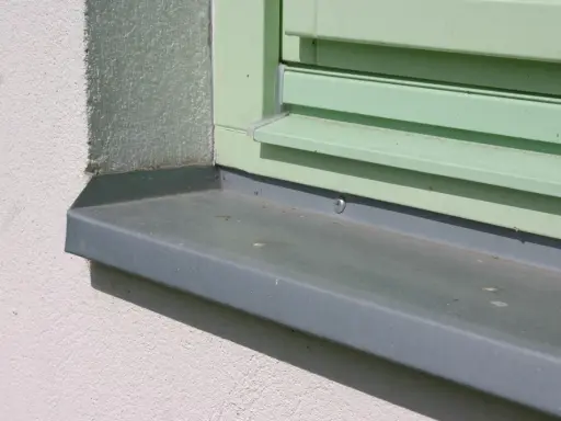 Szary parapet zewnętrzny zamontowany przy oknie z zielonymi okiennicami. Idealny moment, kiedy montować parapety zewnętrzne, to po ociepleniu elewacji.
