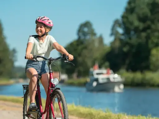 Glad flicka på cykel med hjälm njuter av en cykelsemester med barn längs en kanal. En båt syns i bakgrunden.