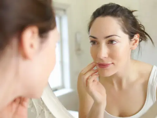 Une femme examine son visage dans un miroir, touchant sa joue. Sa peau semble très fine, peut-être signe d'une carence.