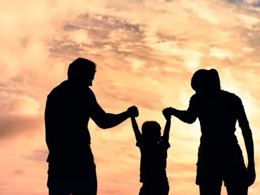Siluetas de una familia jugando bajo un cielo anaranjado. Un niño es levantado por sus padres, un ejemplo de aprendizaje social y emocional.