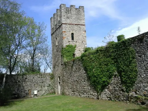 Antica torre del forte di Orino, circondata da mura in pietra e vegetazione rigogliosa, sotto un cielo azzurro.