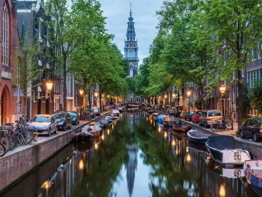 Kanał w Amsterdamie z łodziami, rowerami i wieżą kościelną w tle.