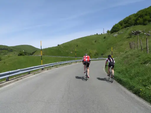 Ciclisti in salita su una strada panoramica, con verdi colline e cielo azzurro. Un'avventura che sembra un **passo cento croci**.