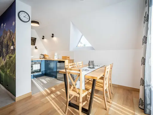 Przytulny apartament pod wierchem. W kuchni niebieskie szafki, jadalnia z drewnianym stołem. Na ścianie fototapeta z górami.