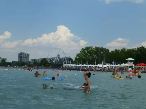 Tłum ludzi cieszy się słońcem i wodą na **Balaton plaża**. W tle widać diabelski młyn i budynki.