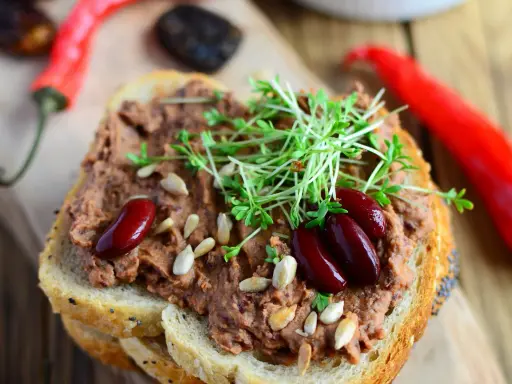 Pyszna pasta z czerwonej fasoli na chlebie, ozdobiona kiełkami i nasionami. Obok leżą czerwone papryczki chili.