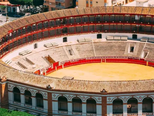 Leere Plaza de Toros mit roten Geländern und sandiger Arena. Die Sitzreihen sind leer, die Sonne scheint auf die traditionelle Architektur.