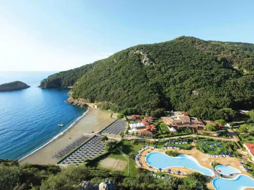 Villaggio toscana sul mare con spiaggia attrezzata, piscine e rigogliosa vegetazione che abbraccia la costa.