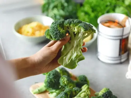 Mano sosteniendo un brócoli fresco, listo para preparar deliciosas recetas de brócoli. En el fondo, queso rallado y zanahorias.