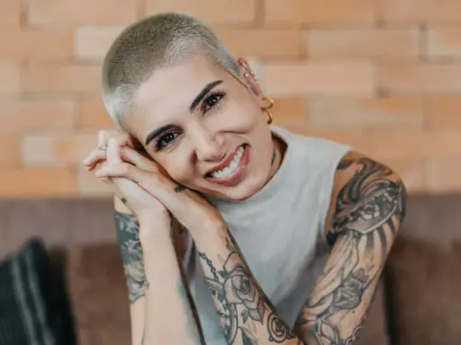 Lächelnde Frau mit Buzz Cut und vielen Tattoos, die ihre Hände vor dem Gesicht verschränkt.