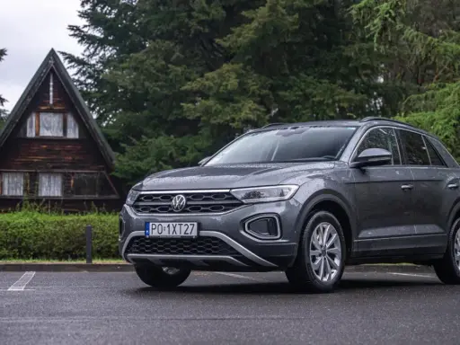 Szary Volkswagen T-Roc używany, zaparkowany przed drewnianym domkiem w lesie. Idealny na wypady za miasto.