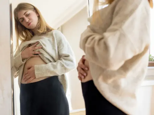 Ragazza si guarda allo specchio, toccandosi la pancia gonfia sopra i pantaloni stretti.