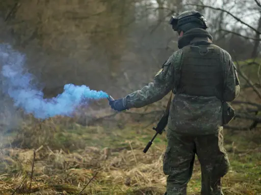Żołnierz w kamuflażu odpala niebieski granat dymny, przestrzegając zasad ASG.