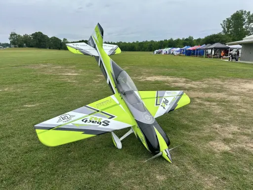 Ein ramy rc-Flugzeug mit auffälligem Design steht auf einer grünen Wiese. Im Hintergrund Zelte und Bäume.