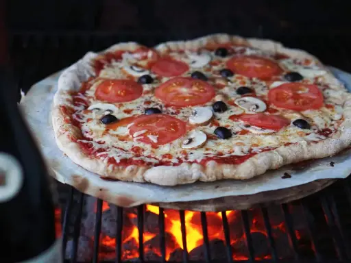 En hemmagjord pizza med tomater, champinjoner och oliver grillas över glödande kol. Perfekt för ett italiensk pizzadeg recept.
