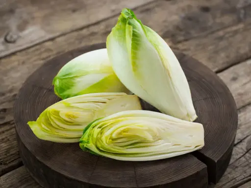 Frische Chicorée-Köpfe, einer davon halbiert, liegen auf einem Holzbrett. Perfekt für einen knackigen Chicorée-Salat.