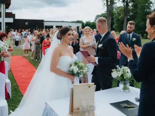 Para młoda podczas ceremonii ślubu cywilnego na zewnątrz. Panna młoda w sukni, pan młody w garniturze. Goście obserwują.