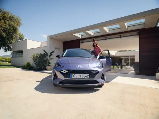 Mujer sonriente junto a un coche alto y morado, aparcado frente a una casa moderna con puertas abiertas.