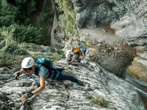 Osoby w kaskach i uprzężach pokonują skalną ścianę, używając liny i karabinków. Cały niezbędny via ferrata sprzęt zapewnia im bezpieczeństwo podczas wspinaczki.
