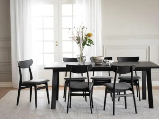 Svart matbord med sex stolar, en vas med blommor och en skål. Modern inredning matplats med ljus matta och vita dörrar i bakgrunden.