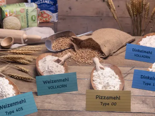 Verschiedene Mehlsorten, darunter Weizenmehl, Dinkelmehl und Pizzamehl, liegen in Holzschalen. Daneben Weizenkörner und ein Nudelholz.