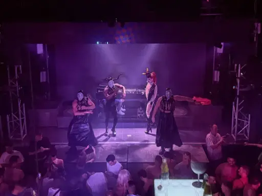 Performers in masks and elaborate outfits dance on stage at one of Valencia's vibrant gay bars, while a crowd watches below.