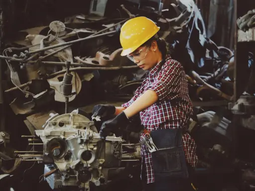 Mujer con casco amarillo y gafas de seguridad trabajando en un motor. Usa guantes negros y un delantal de mezclilla, demostrando el uso de los equipos de protección individual.