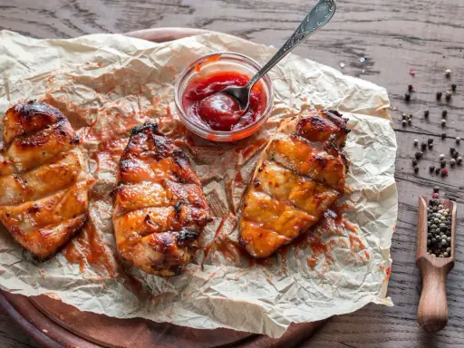 Saftiges Grillfleisch zart machen: Drei gegrillte Hähnchenbrustfilets mit Sauce auf Backpapier, daneben eine Schale Ketchup und eine Holzschaufel mit Pfefferkörnern.