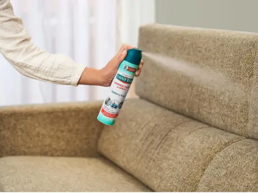 Mano rociando desinfectante en spray sobre un sillón de tela. Una forma fácil de saber como limpiar sillones de tela y eliminar olores.