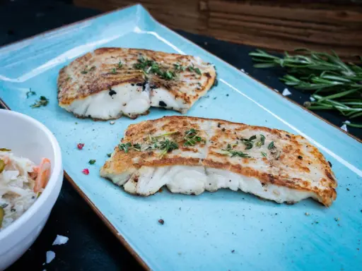 Zwei perfekt gegrillte Kabeljaufilets, mit Kräutern bestreut, serviert auf einem blauen Teller. Genießen Sie kabeljau grillen ohne alufolie!