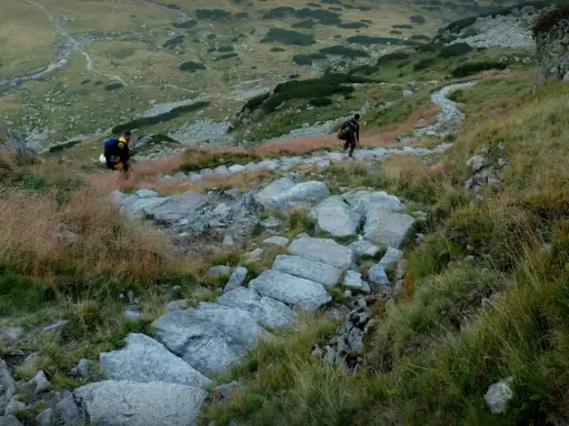 Dwóch turystów z plecakami przemierza kamieniste szlaki z Kuźnic, podziwiając górski krajobraz.
