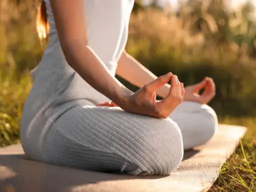 En person sitter i lotusställning på en matta utomhus, med händerna i meditation. Solen lyser genom gräset och skapar en varm atmosfär för morgonmeditation.